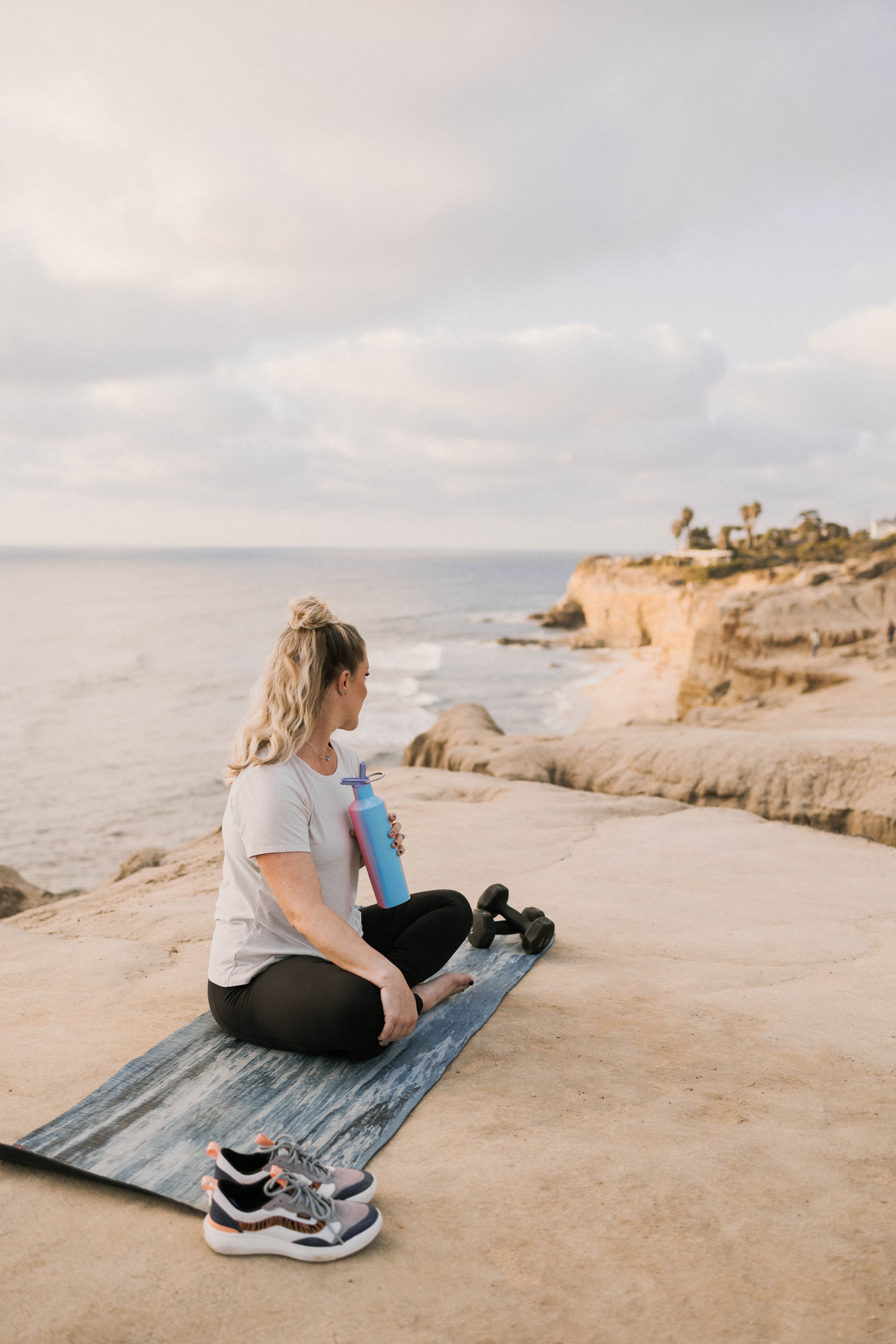 Woman sitting on a yoga mat on a beach, with shoes nearby, looking at the ocean under cloudy sky.