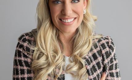 Woman with long blonde hair smiling, wearing a patterned blazer and white top, arms crossed, neutral background.