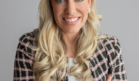 Woman with long blonde hair smiling, wearing a patterned blazer and white top, arms crossed, neutral background.