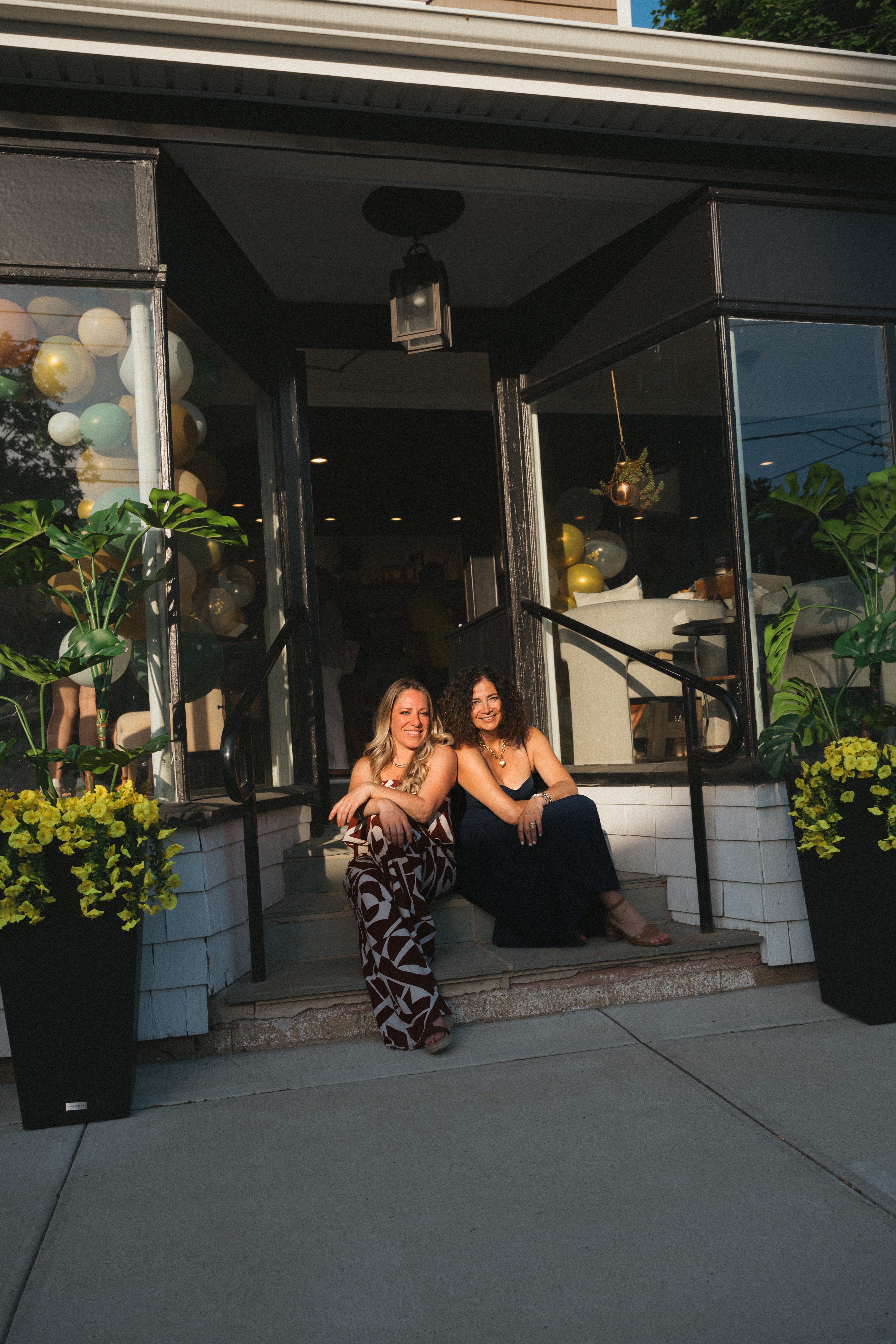 Two women sit on steps outside a building entrance, surrounded by potted plants and balloons, during daytime.