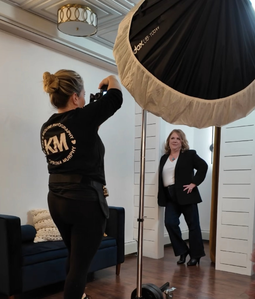 Photographer taking picture of woman in black blazer and white shirt in a room with white walls and a navy sofa.
