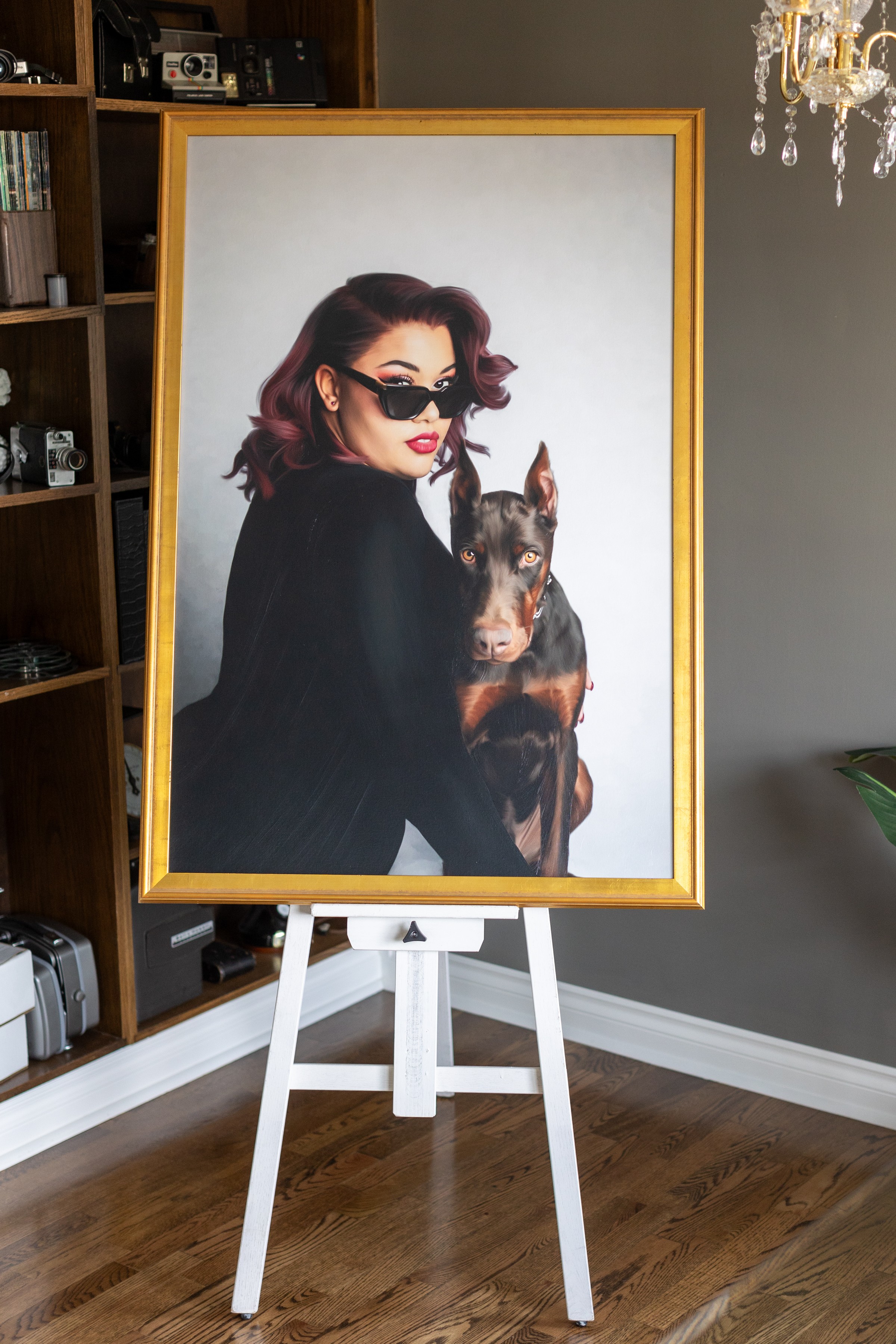 Portrait of a woman with dark hair and sunglasses holding a dog, framed and displayed on an easel.