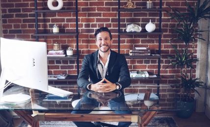 Man in suit sitting at glass desk with computer, brick wall and shelves behind him, smiling.
