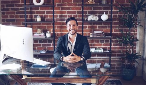 Man in suit sitting at glass desk with computer, brick wall and shelves behind him, smiling.