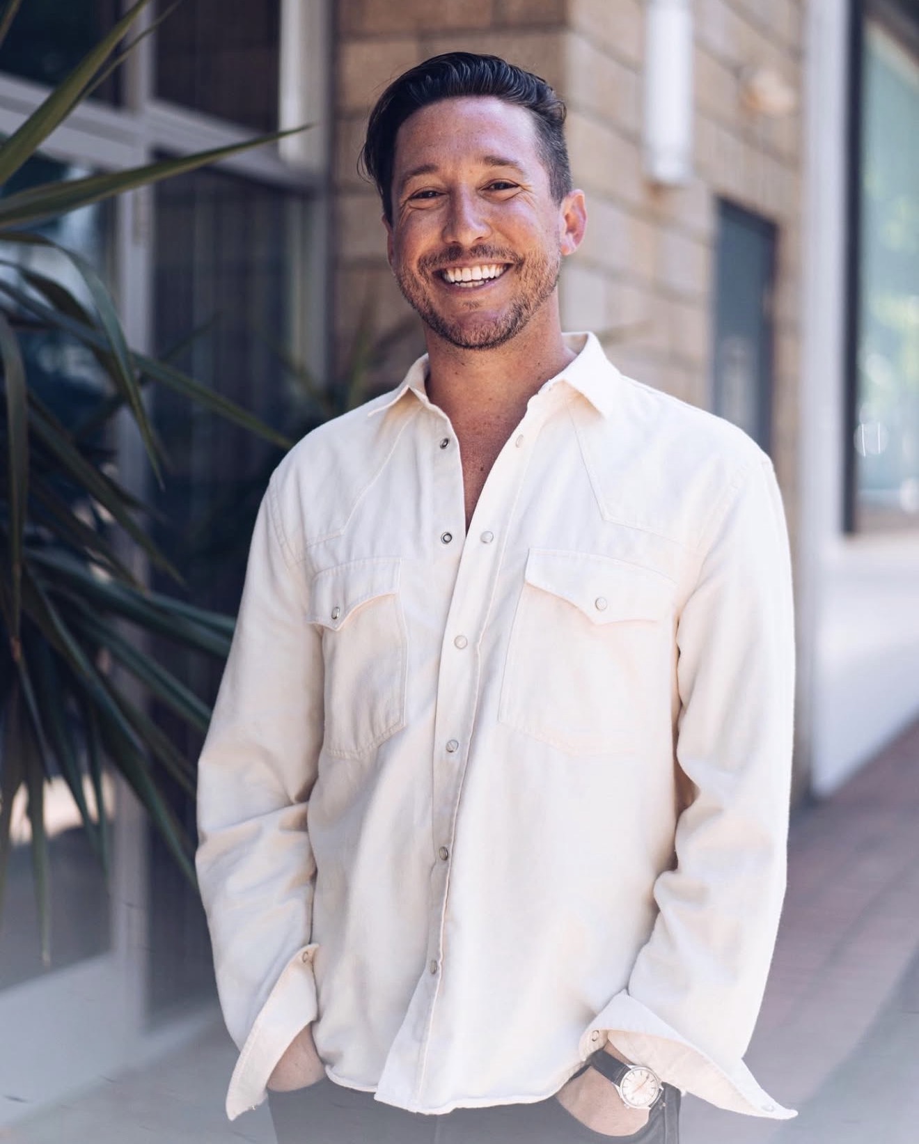 Smiling man in a white shirt standing outdoors in front of a building with plants nearby.