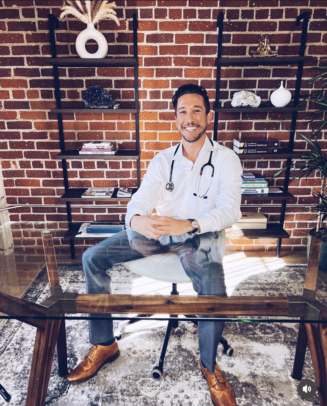 Doctor sitting at desk with brick wall and shelves behind, smiling, wearing white coat and stethoscope.