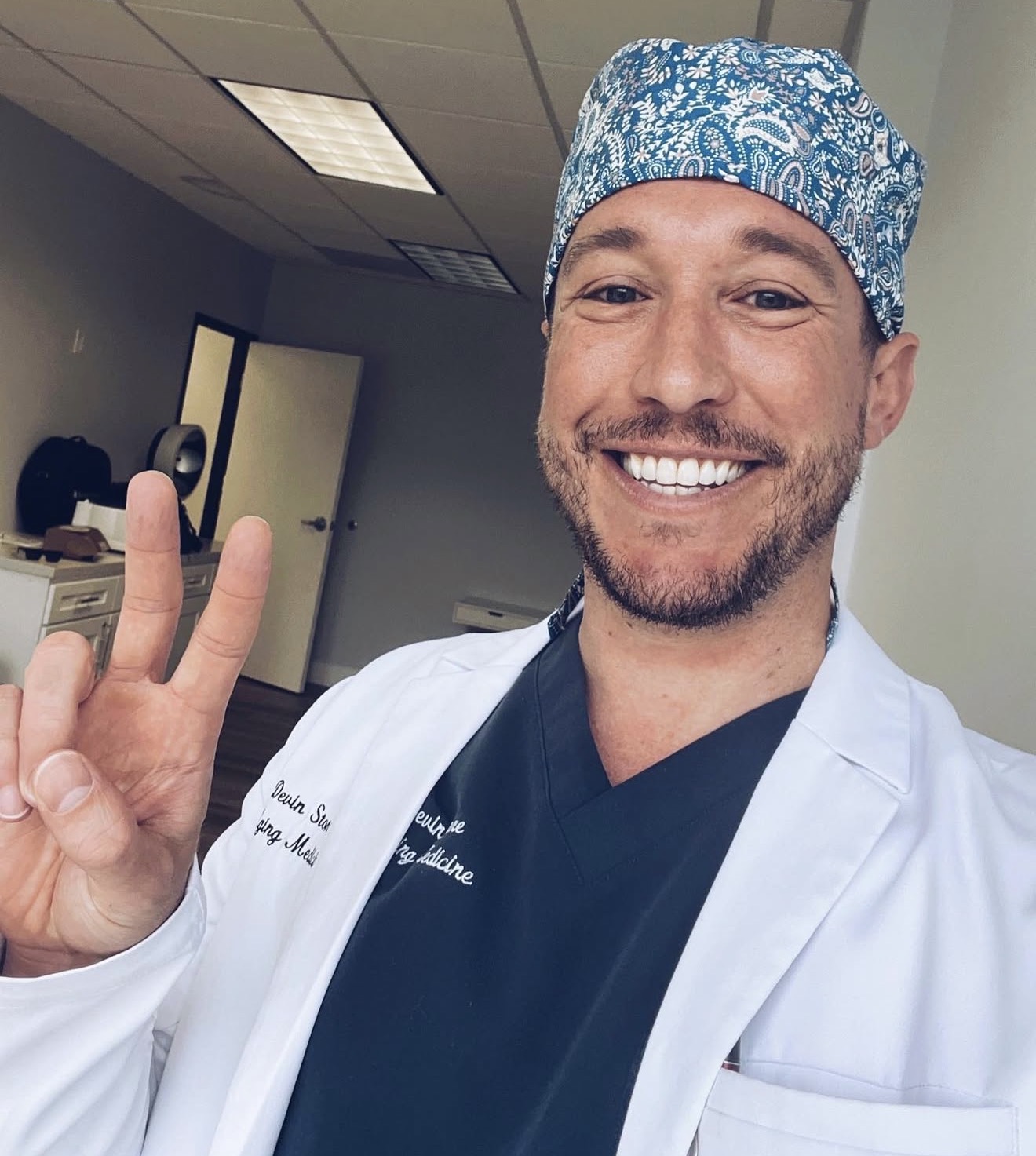 Smiling man in medical attire making a peace sign, wearing a patterned surgical cap, in a clinical setting.