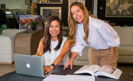 Two women working together at a table with a laptop and open books in a modern office or lounge area.