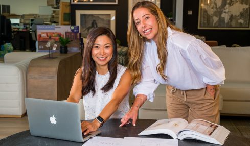 Two women working together at a table with a laptop and open books in a modern office or lounge area.