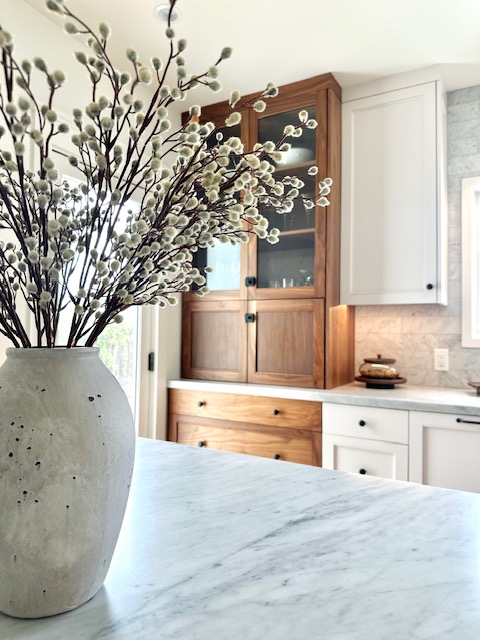 Close-up of a vase with white flowering branches on a kitchen countertop, with cabinets and a window in the background.