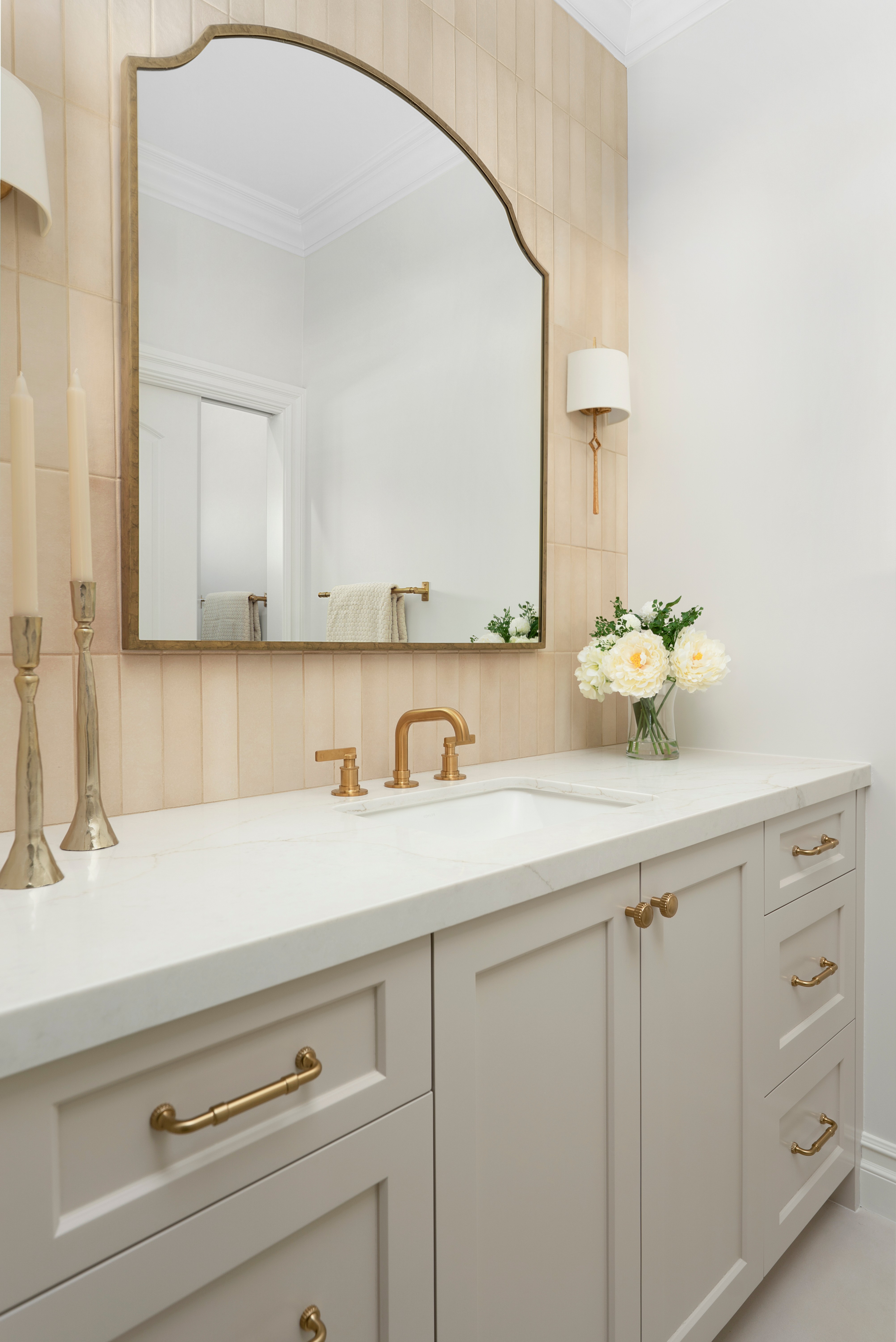 Bathroom vanity with a large mirror, white countertop, gold fixtures, and a vase of white flowers, against a light wood wall.