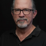 Portrait of a middle-aged man with glasses, gray hair, and a beard, wearing a black polo shirt, against a dark background.