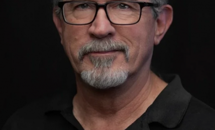 Portrait of a middle-aged man with glasses, gray hair, and a beard, wearing a black polo shirt, against a dark background.
