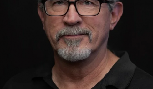 Portrait of a middle-aged man with glasses, gray hair, and a beard, wearing a black polo shirt, against a dark background.