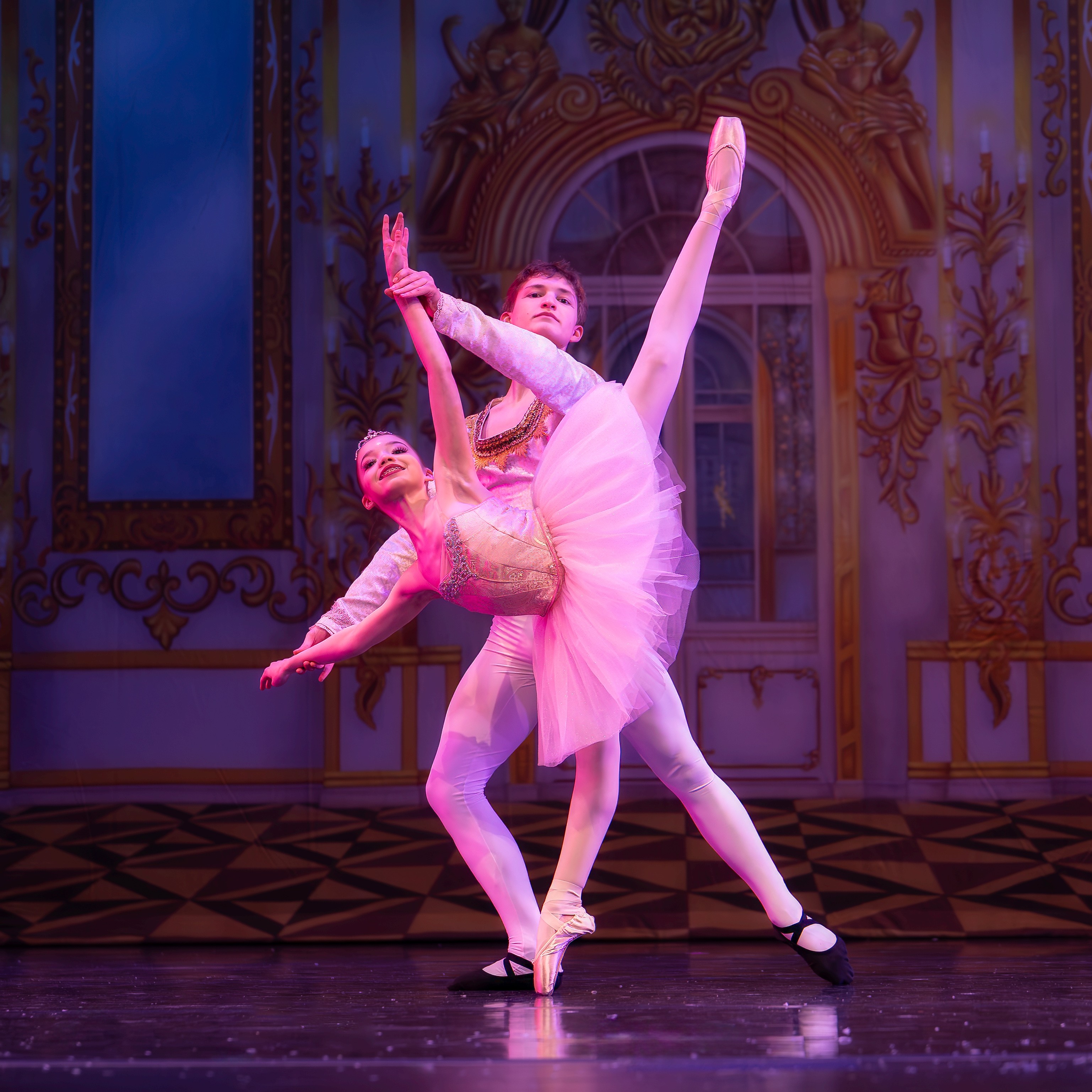 Two ballet dancers perform on stage with ornate background, one lifting the other in a pose, both in pink costumes.