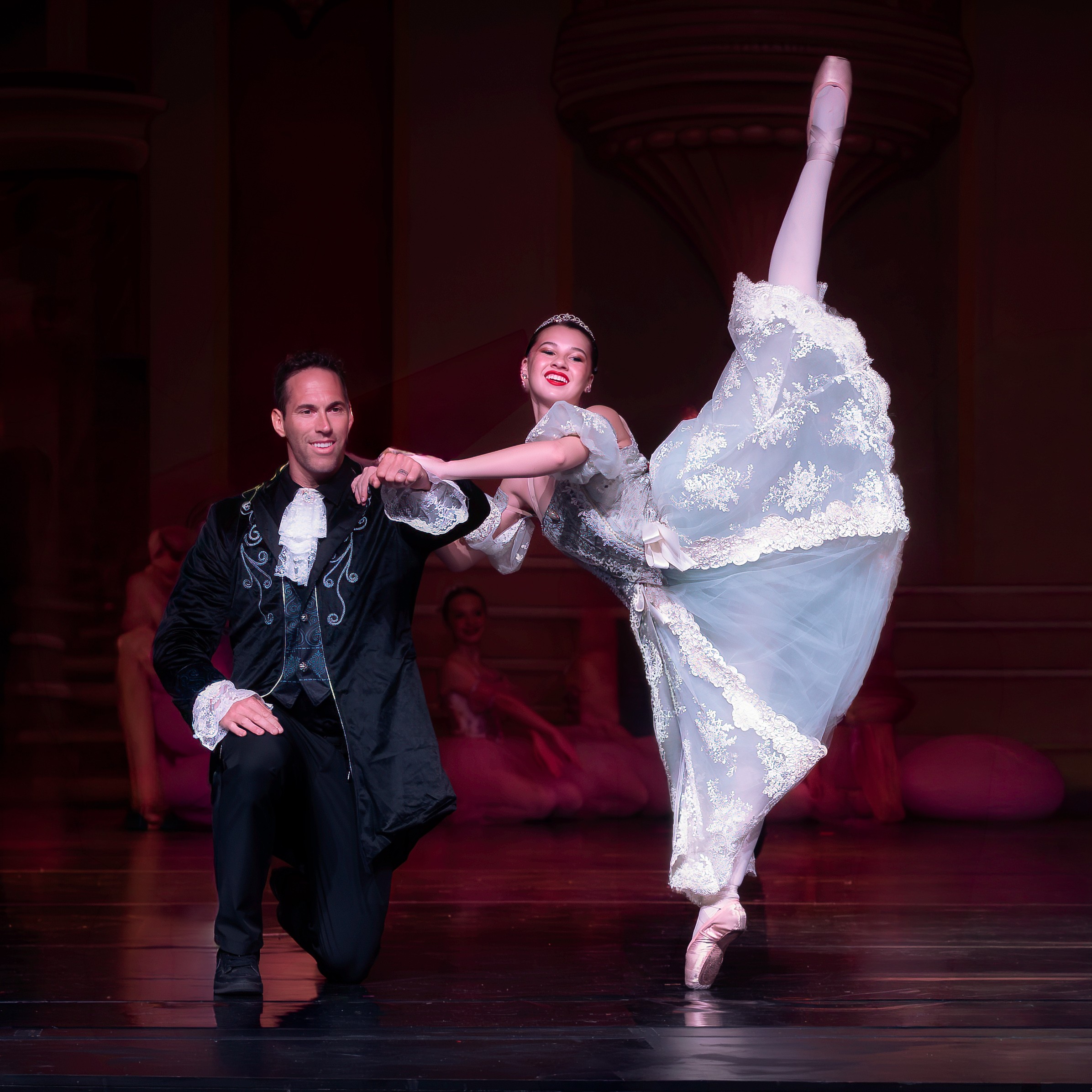 Ballet dancers perform on stage, one standing and the other executing a high leg extension, with a dark background.