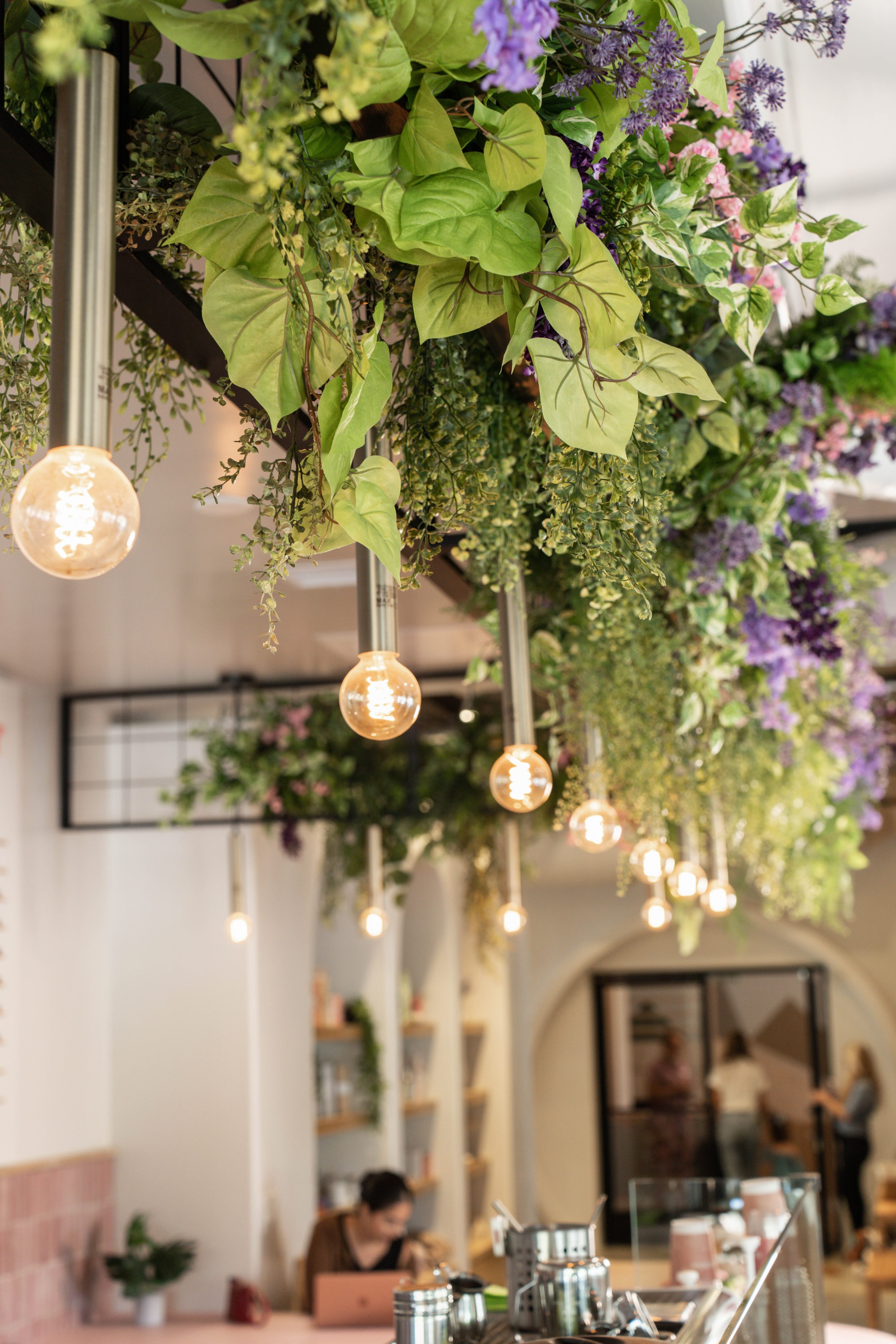 Hanging floral arrangement with purple and green flowers and leaves, illuminated by hanging light bulbs, inside a cafe or restaurant.