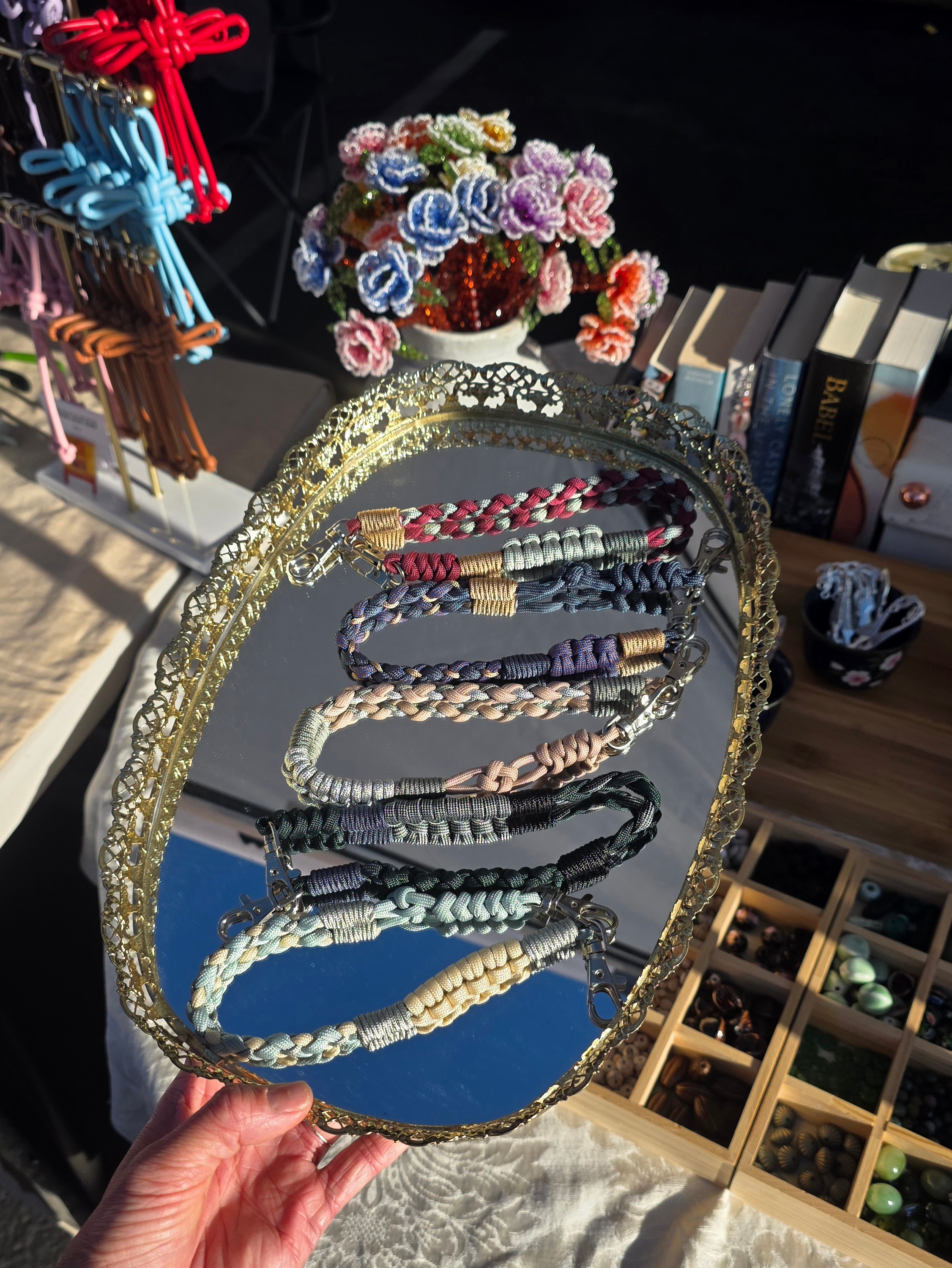 Hand holding a tray with five beaded necklaces, a vase of colorful flowers, and books on a table, outdoors.