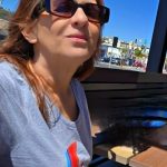 Woman with sunglasses sitting outdoors at a table, with buildings and cars in the background.