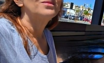Woman with sunglasses sitting outdoors at a table, with buildings and cars in the background.