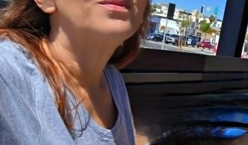 Woman with sunglasses sitting outdoors at a table, with buildings and cars in the background.