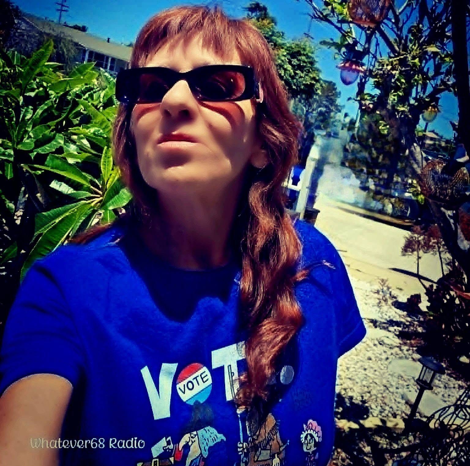 Woman with sunglasses outdoors, wearing a blue shirt, surrounded by plants and trees, sunny day.