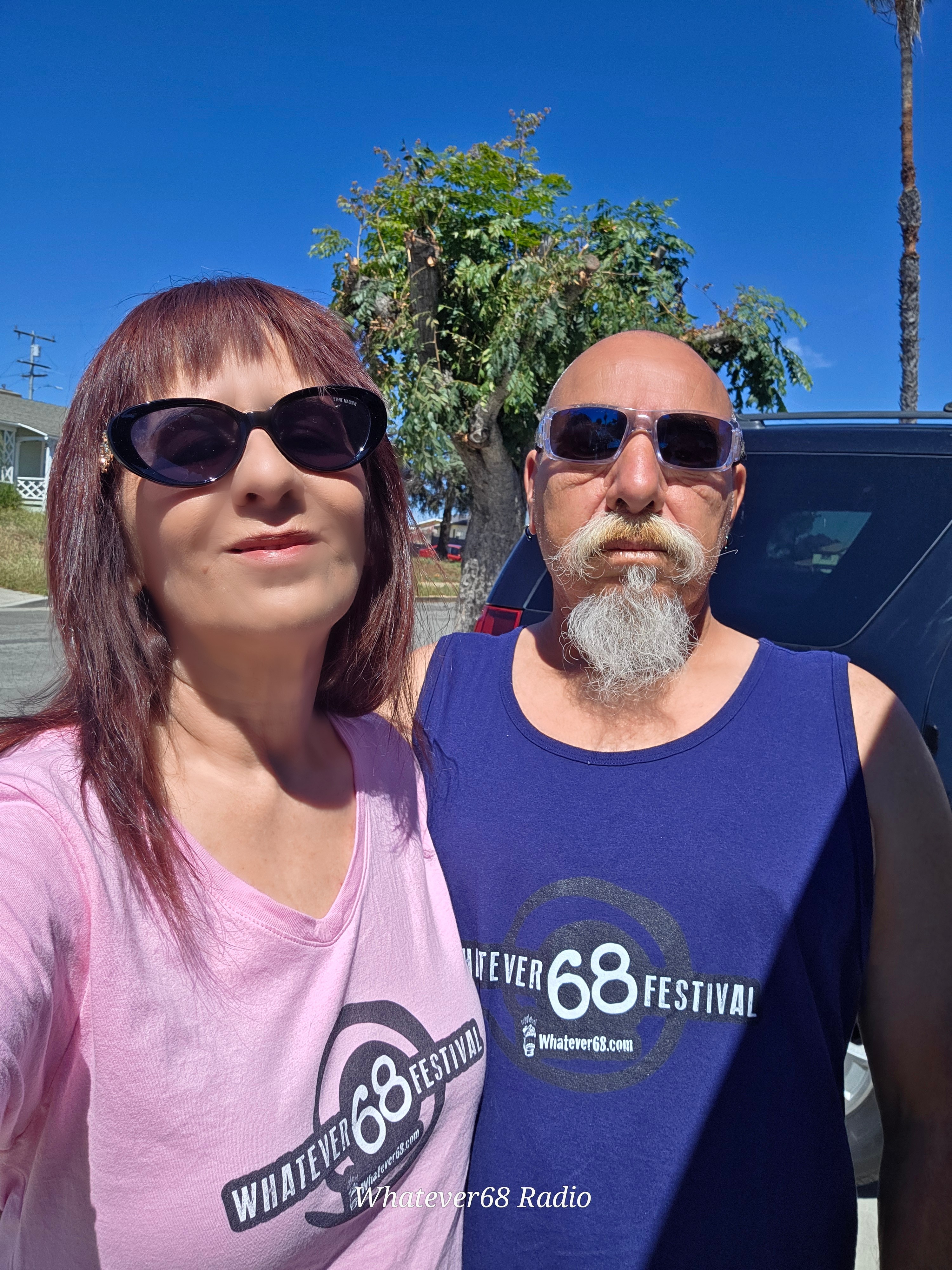 Two people wearing sunglasses outdoors, standing in front of a tree and a vehicle, sunny day with clear blue sky.
