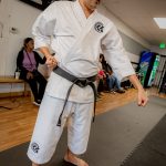 Martial artist in white gi with black belt practicing in a dojo, with people seated in the background.