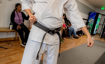 Martial artist in white gi with black belt practicing in a dojo, with people seated in the background.