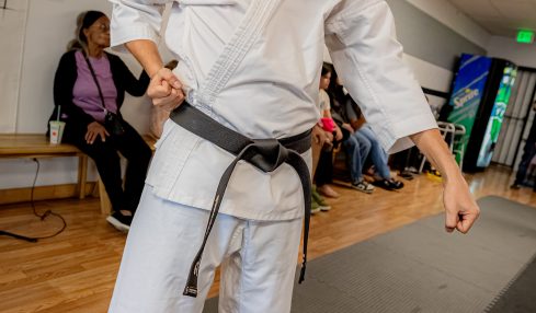 Martial artist in white gi with black belt practicing in a dojo, with people seated in the background.