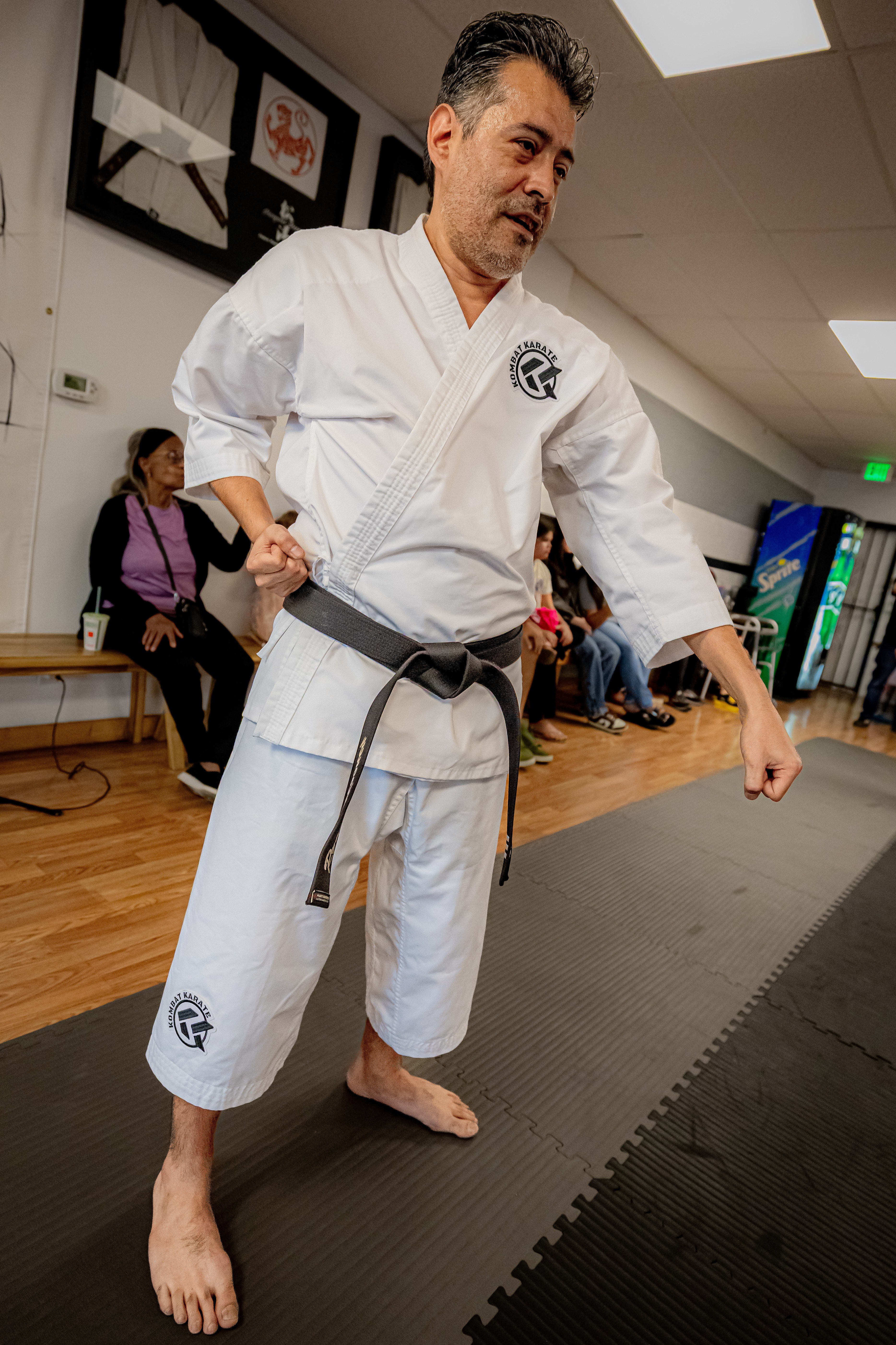 Martial artist in white gi with black belt practicing in a dojo, with people seated in the background.