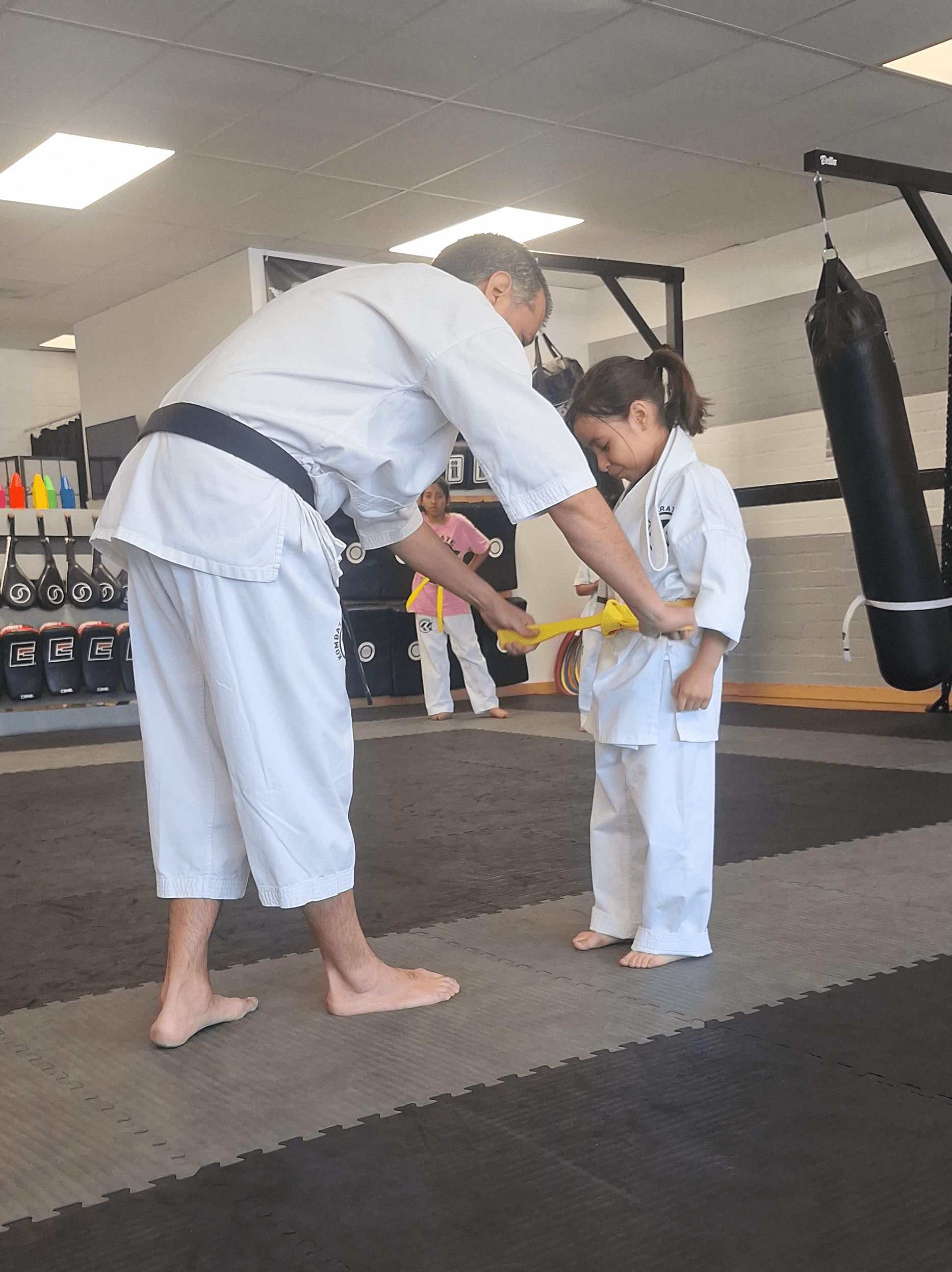 Instructor teaching a young girl in martial arts, both wearing white uniforms, in a gym with punching bags.