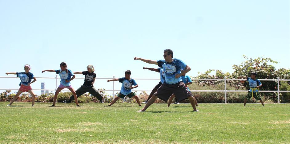 Group of children practicing martial arts outdoors on grass field, with a fence and trees in background.