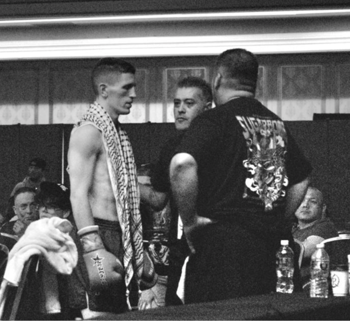 Three men engaged in conversation, one shirtless with boxing gloves, in a crowded indoor setting with bottles on a table.