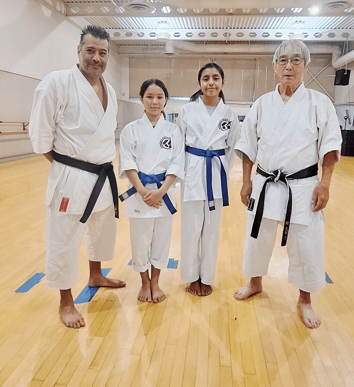 Four people in martial arts uniforms standing on a wooden floor in a dojo, two adults and two children, all barefoot.