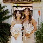 Two women in white dresses holding bouquets, smiling outdoors with greenery and a white wall background.