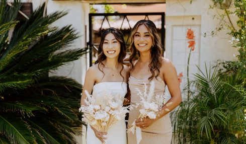 Two women in white dresses holding bouquets, smiling outdoors with greenery and a white wall background.