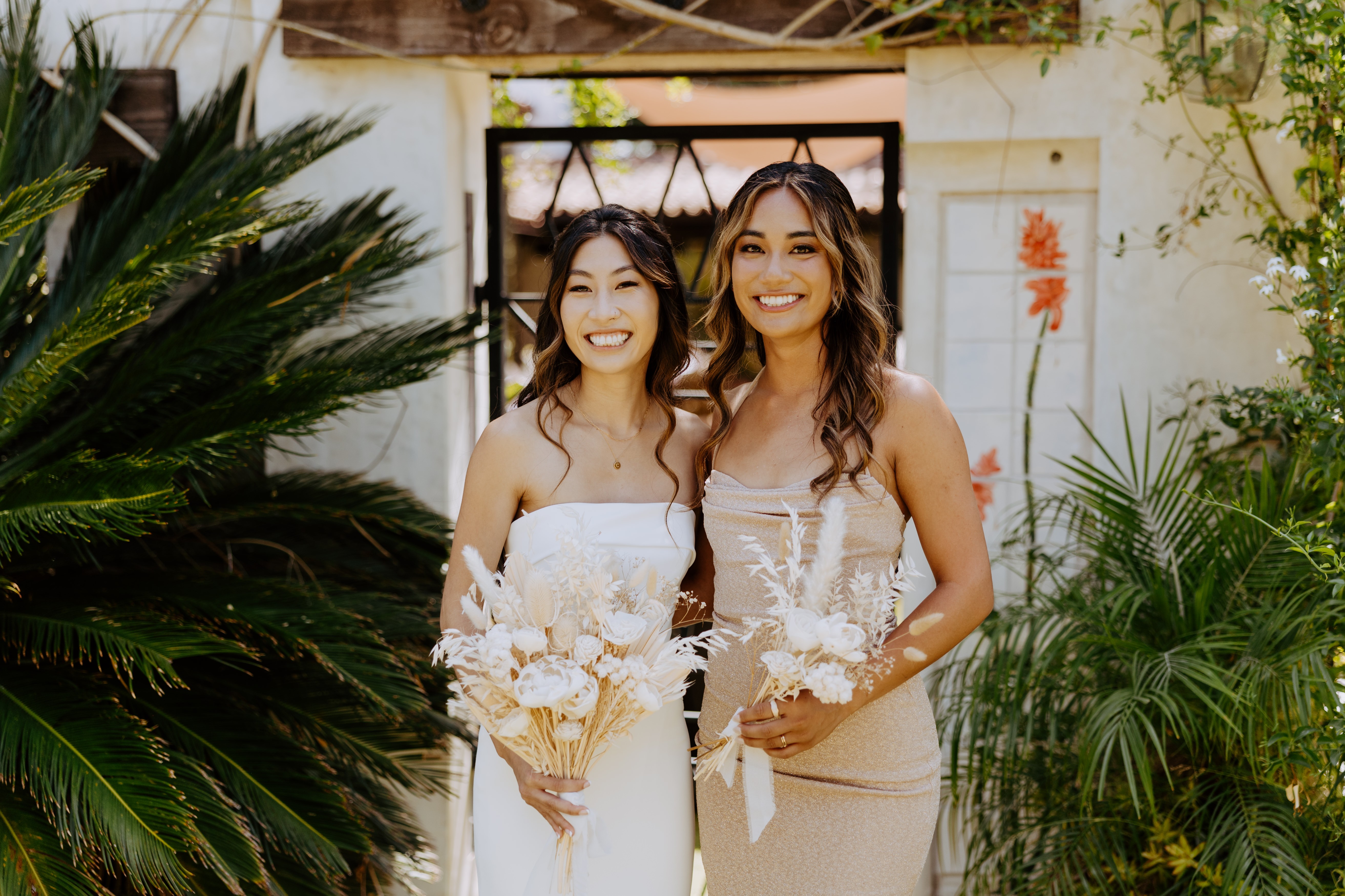 Two women in white dresses holding bouquets, smiling outdoors with greenery and a white wall background.