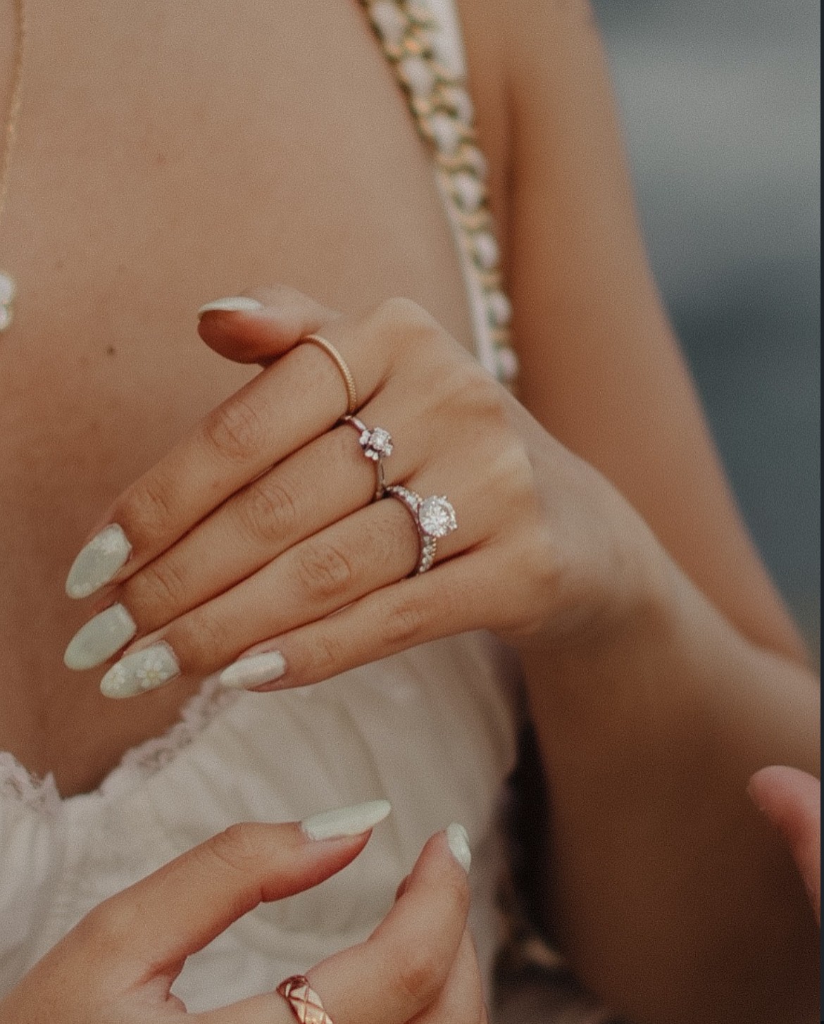 Close-up of a woman's hand with rings, showing manicured nails and jewelry, against a blurred background.