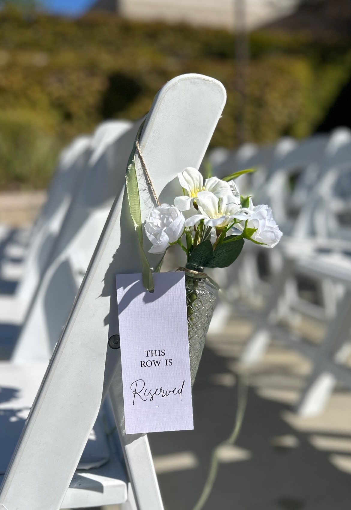 White chair with a bouquet of white flowers attached, and a reserved sign hanging on the backrest, outdoors with chairs in background.