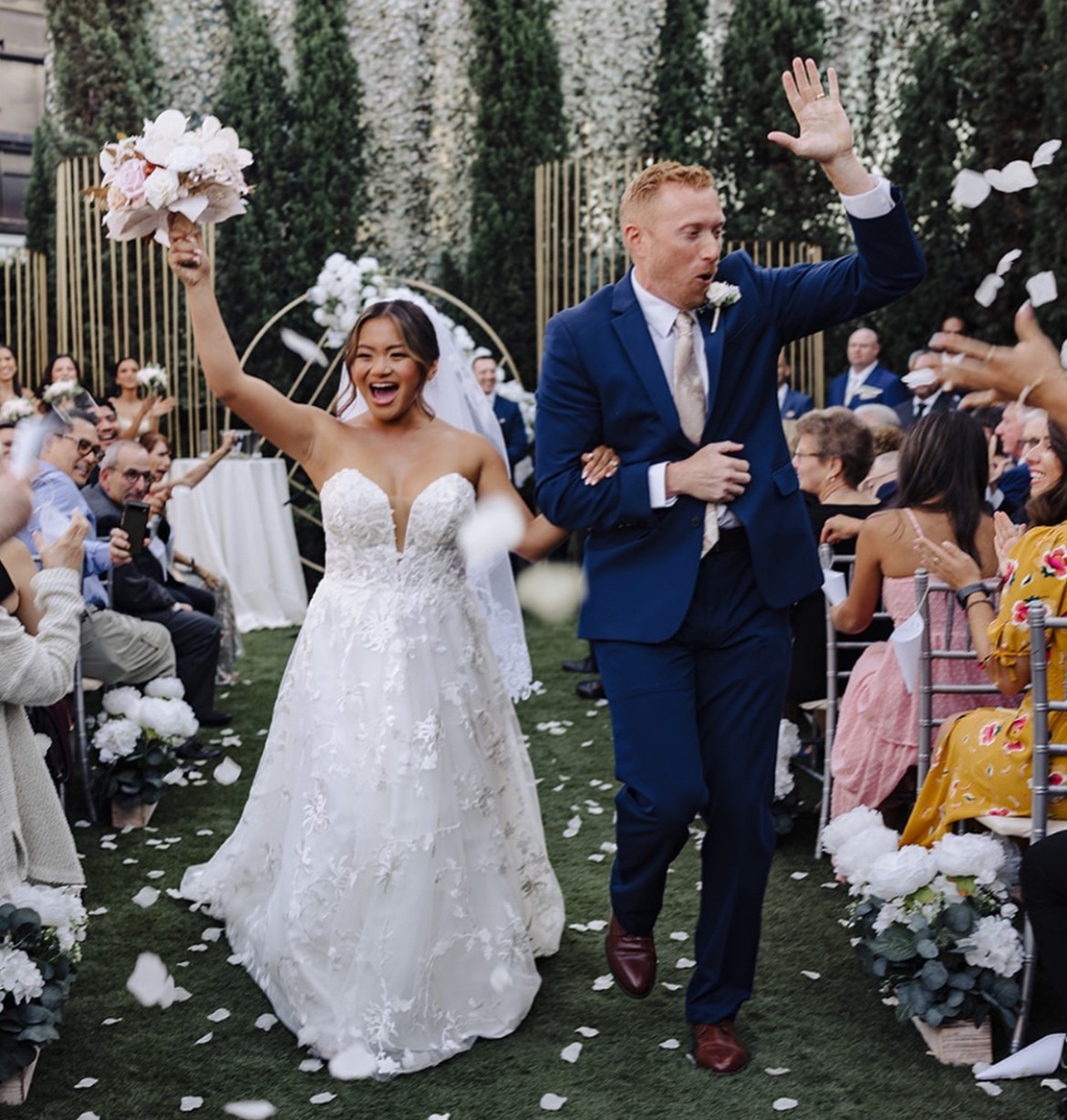 Bride and groom walking down aisle, smiling, surrounded by seated guests, outdoor wedding with floral decorations, bride holding bouquet, groom in blue suit.