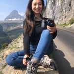 Smiling woman sitting on a rock by a mountain road, holding a camera, with mountains and cliffs in background.