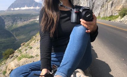 Smiling woman sitting on a rock by a mountain road, holding a camera, with mountains and cliffs in background.