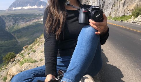 Smiling woman sitting on a rock by a mountain road, holding a camera, with mountains and cliffs in background.