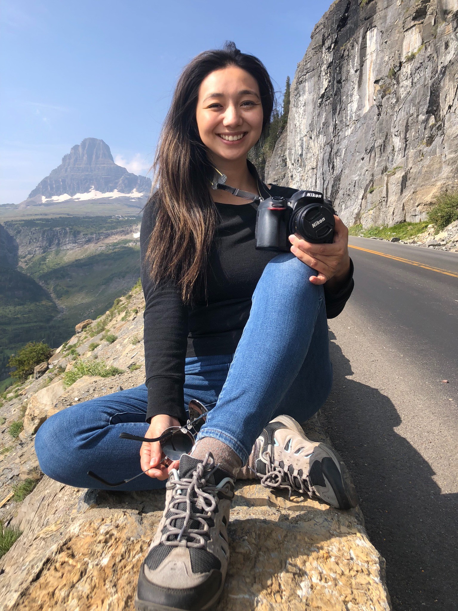 Smiling woman sitting on a rock by a mountain road, holding a camera, with mountains and cliffs in background.