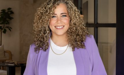 Smiling woman with curly hair wearing a purple blazer and white top, standing indoors near a black door frame.