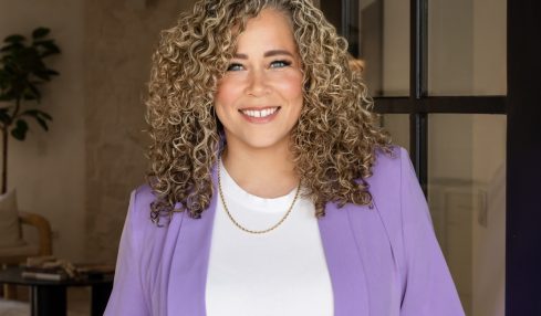 Smiling woman with curly hair wearing a purple blazer and white top, standing indoors near a black door frame.