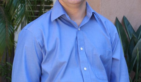Smiling man in a blue shirt standing outdoors with plants in background.