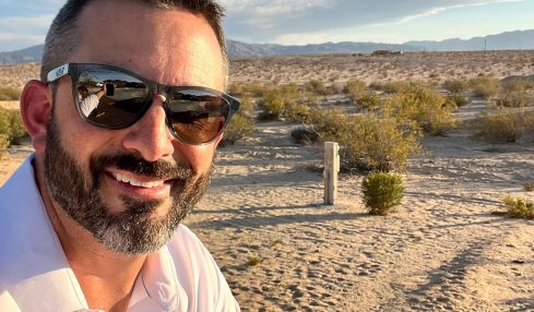 Man with sunglasses and beard taking selfie outdoors on sandy terrain with blue sky and clouds.