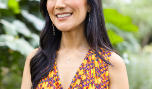 Smiling woman with long dark hair in a colorful sleeveless dress outdoors, greenery in background.
